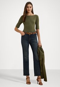 Olijfgroene aangesloten top met driekwartmouwen, gecombineerd met donkerblauwe rechte jeans en een bruine riem. Houdt een gestructureerde groene jas vast.