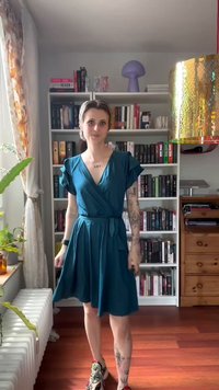 Pessoa com cabelo curto, vestindo um vestido teal e ténis, está em frente a uma estante de livros numa sala com chão de madeira e plantas perto da janela.