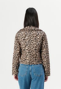 Kvinna med rakt svart hår, iförd en leopardmönstrad jacka och blå jeans, stående mot en vit bakgrund.