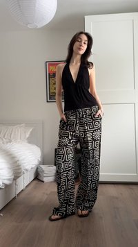 Νεαρή γυναίκα που στέκεται μέσα σε εσωτερικό χώρο, φορούσα μαύρο τοπ halter και φαρδιά παντελόνια με σχέδιο σε ασπρόμαυρους τόνους, καθώς και σανδάλια.