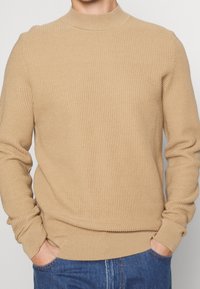 Beige Rippenstrickpullover mit hohem Rundhalsausschnitt und langen Ärmeln. Der Stoff hat eine strukturierte Oberfläche und wird mit blauen Jeans kombiniert.