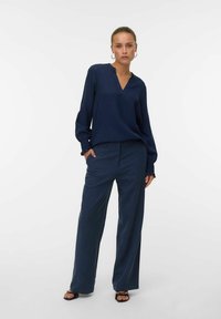 Mörkblå blus med långa ärmar, V-ringning och texturerad tyg, parat med vida mörkblå byxor. Enkel design med manschettdetaljer.