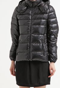 Black puffer jacket with a shiny finish, zippered front, and two side pockets. Features a hood and a quilted design with puffed sections.