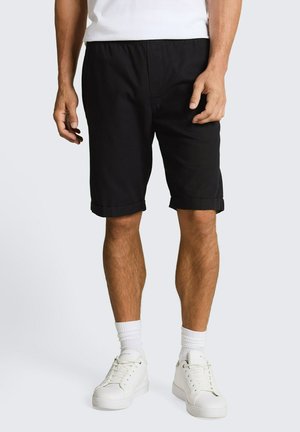 Mann trägt schwarze Shorts mit umgeschlagenem Saum, weiße Socken, weiße Turnschuhe und ein weißes Hemd, steht vor einem schlichten hellen Hintergrund.