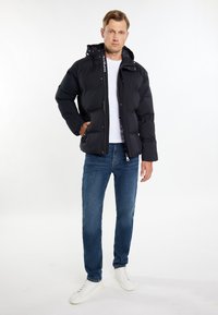 Sort pufferjakke med hætte, lynlås foran og trykknaplukning. Har en ensartet tekstur og logodetalje på lynlåsens træk. Bæres med jeans.