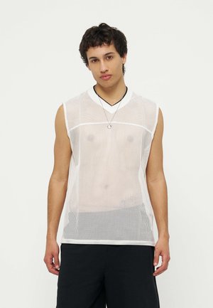 Jonge man draagt een mouwloos wit mesh topje over een zwart onderhemd, met zwarte broek en een ketting, staand tegen een effen achtergrond.