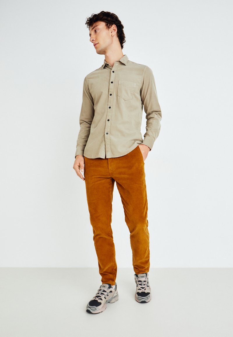 Chemise boutonnée marron clair avec une poche, associée à un pantalon en velours côtelé couleur rouille et des baskets grises avec des accents noirs.