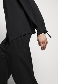 Chemise noire à manches longues, coupe légèrement oversize, fentes latérales et poignets noués, associée à un pantalon noir ample assorti. Texture lisse.