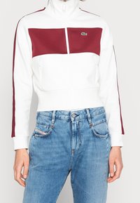 Person som bär en vit zip-up jacka med vinröda paneler och ränder, i kombination med hög midja ljusblå jeans.