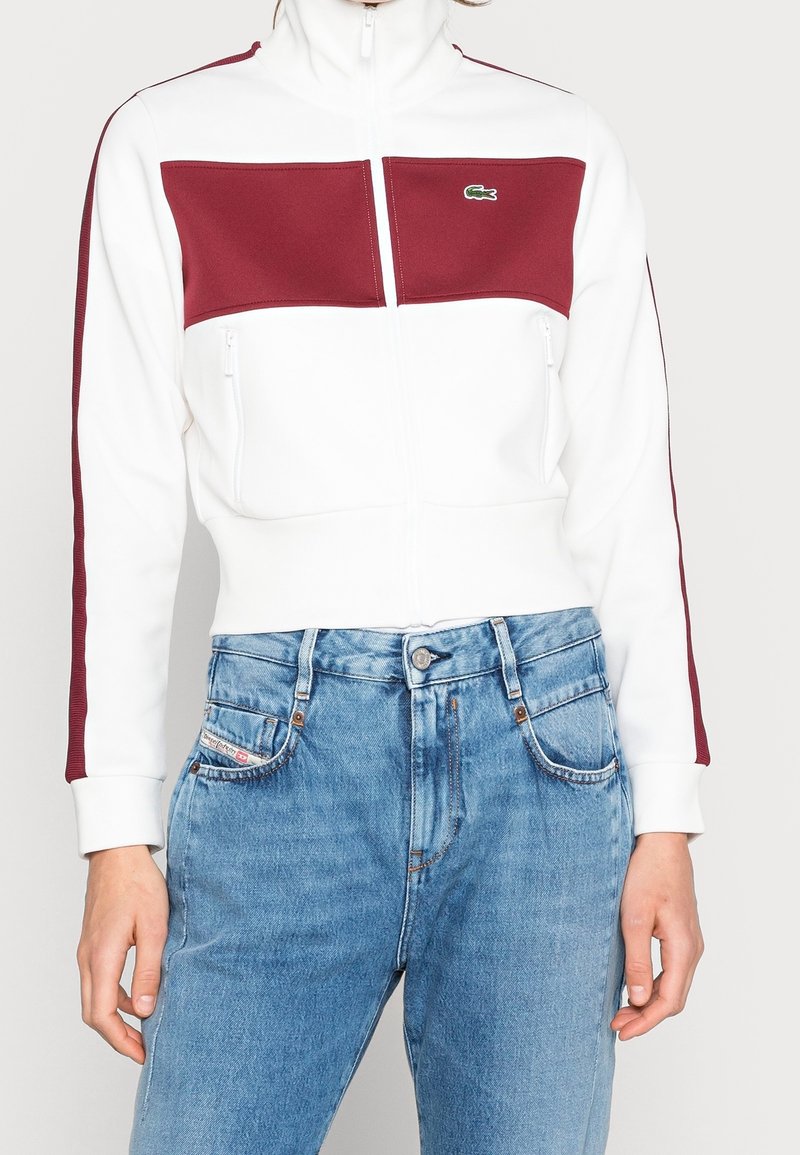 Person som bär en vit zip-up jacka med vinröda paneler och ränder, i kombination med hög midja ljusblå jeans.