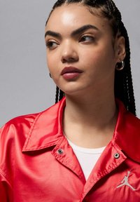 Punainen satiinitakki, jossa on terävä kaulus, hopeiset yksityiskohdat ja Jordan-logo. Päällä valkoinen ribbineulottu paita.