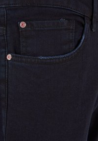 Tumedad denim püksid on varustatud tagataskuga, millel on punased metallist neetid, nähtav õmblus ja sile tekstuur, peegeldades sügavat sinise-musta tooni.