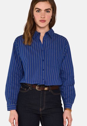 Femme portant une chemise bleue à manches longues à rayures blanches verticales, rentrée dans un jean foncé avec une ceinture marron.