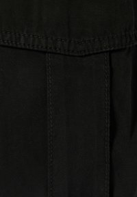 Lähikuva mustasta kankaasta, jossa näkyy teksturoidun pinnan ja ompeleiden yksityiskohtia. Sisältää taskun ja yläreunassa olevan taitteen.