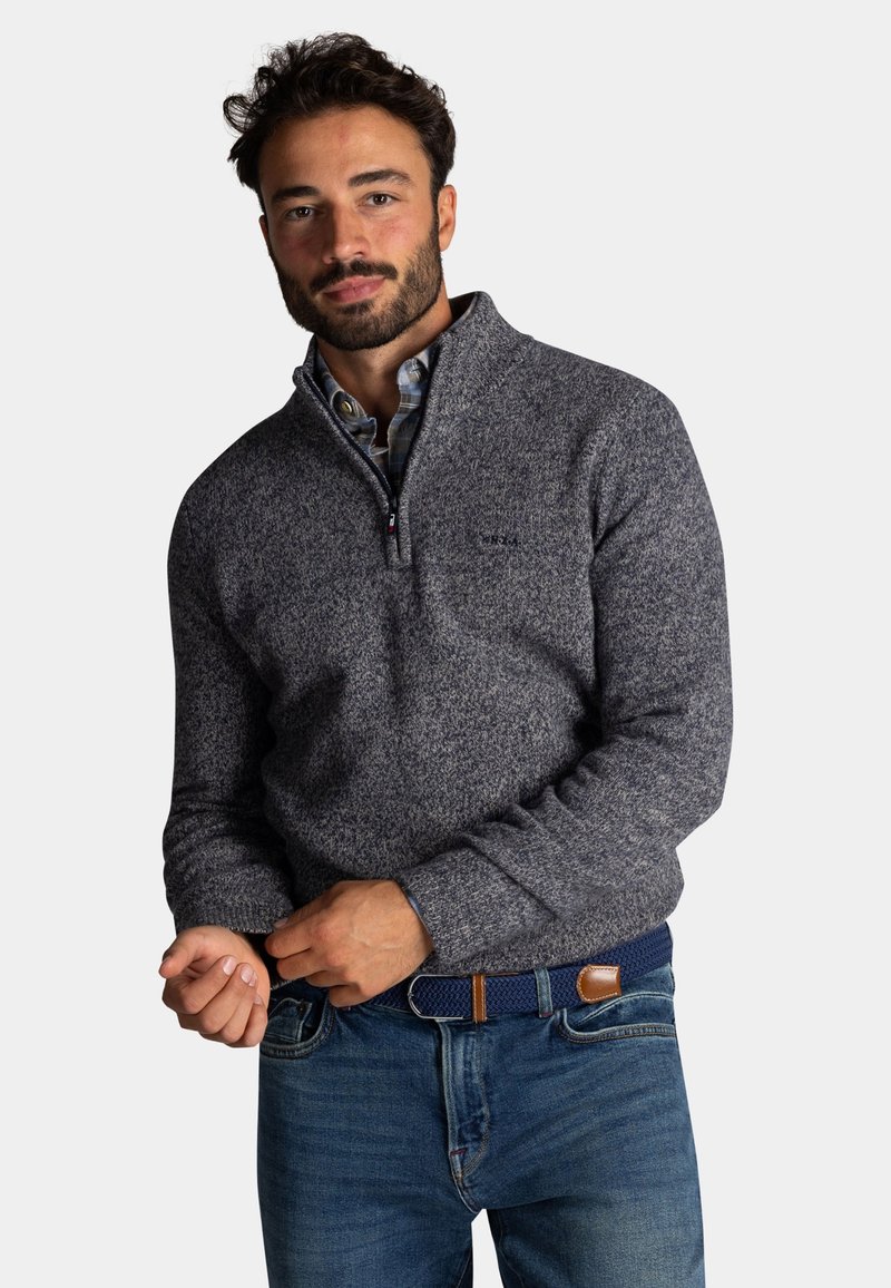 Grijze gebreide trui met een ritskraag, gemaakt van een gestructureerde stof en met ribbels aan de mouwen, gedragen over een geruite blouse met blauwe spijkerbroek.