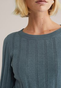 Maglione lavorato a maglia in un verde bluastro tenue, con costine verticali e un classico scollo rotondo. Il tessuto presenta una finitura morbida e testurizzata.
