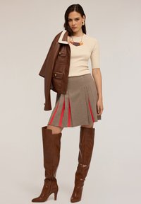 Femeie îmbrăcată cu un top bej și o fustă cu modele și pliuri roșii, ținând o jachetă din piele maro pe umăr, purtând cizme maro până la genunchi și un colier.
