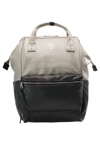 Sac à dos bicolore avec une partie supérieure beige clair et une grande poche avant zippée noire, poignées supérieures et fermetures éclair latérales.