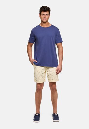 Joven de pie vistiendo una camiseta azul de manga corta, pantalones cortos beige con estampados de palmeras y zapatos casuales azul marino.