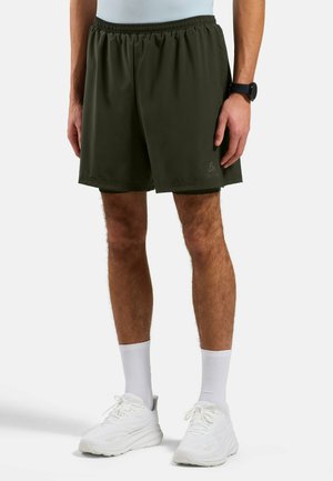 Mann trägt dunkelgrüne Sportshorts, weiße Socken und weiße Turnschuhe, steht vor einem schlichten hellen Hintergrund.