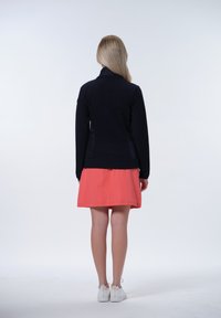 Marinblå zip-up jacka med strukturerade sidopaneler, i kombination med en korallfärgad kjol. Modellen bär vita sneakers med guldiga dragkedjor bak.
