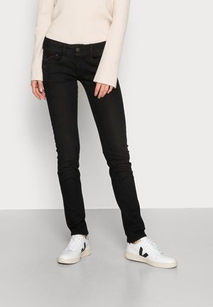 Frau trägt schwarze Slim-Fit-Jeans, weißes geripptes Langarmshirt und weiße Sneaker mit schwarzem Logo, steht leicht mit gekreuzten Beinen.