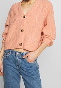 Kort cardigan i mjukt, texturerat rosa material med djup v-ringning, tre stora bruna knappar och ribbad fåll, i kombination med jeans.