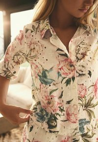 Blommig blus i vit väv med broderade detaljer och utskurna mönster. Har korta ärmar, krage och knappar.