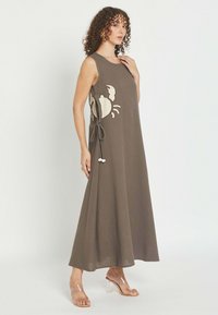 Mujer con vestido largo marrón sin mangas con diseño grande de cangrejo blanco y detalle de lazo lateral, posando frente a fondo liso y claro.