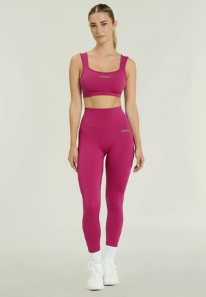 Nainen seisoo päällään kirkkaanpinkki urheilurintaliivit ja korkeavyötäröiset leggingsit valkoisilla sukilla ja tennareilla yksivärisellä taustalla.