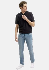 Czarna koszulka polo z krótkim rękawem i zamkiem w kołnierzu, w połączeniu z jasnoniebieskimi dżinsami o wąskim kroju i białymi trampkami. Model stoi w neutralnej pozycji.