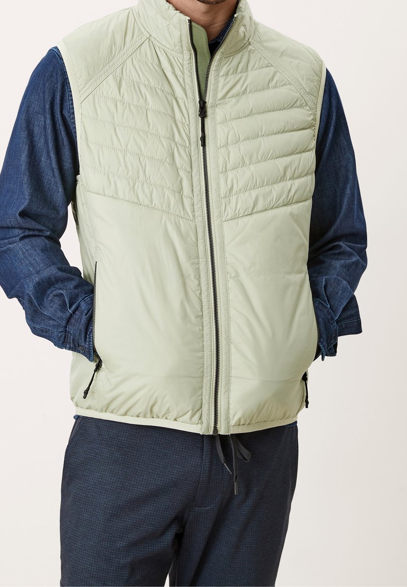 Homme portant une veste matelassée vert clair sur une chemise bleue à manches longues, les mains dans les poches de la veste, sur un fond uni.