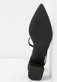 Chaussure à talon haut noire à bout pointu avec des brides transparentes reposant sur son côté sur un fond blanc.