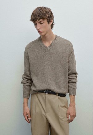 V-neck sweater in light brown textured knit, with long sleeves and ribbed cuffs, paired with beige trousers and a dark patterned belt.