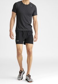 Mies, jolla on yllään musta teksturoitu lyhythihainen urheilupaita, mustat Under Armour -shortsit ja harmaat juoksukengät valkoista taustaa vasten.