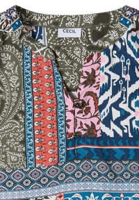 Blouse multicolore col avec des motifs patchwork en vert, bleu, rose et rouge, présentant des dessins floraux et géométriques et deux boutons sous le col.