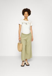 Camiseta branca com gráfico "PALM PARADISE", calças largas verde claro, sandálias estampadas e uma bolsa de palha com detalhes de contas.