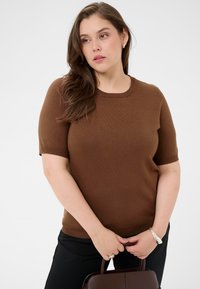 Pull en maille marron à manches courtes avec un col rond, associé à un pantalon noir. Les accessoires comprennent un bracelet en argent et un sac à main marron.