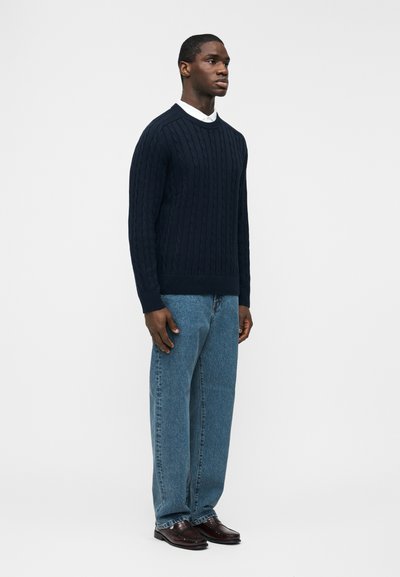 Άνδρας που στέκεται φορώντας πουλόβερ navy με πλέξη καλώδιο, λευκό πουκάμισο με γιακά από κάτω, άνετο μπλε τζιν και σκούρα καφέ δερμάτινα παπούτσια σε απλό φόντο.