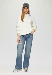 Witte hoodie, blauwe versleten jeans met scheuren, en zilveren sportchoenen. Model draagt een grijze pet en staat tegen een neutrale achtergrond.