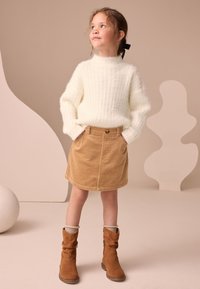 Pull en maille crème, jupe en velours côtelé couleur noisette avec poches avant et bouton, bottes marron décontractées, chaussettes gris clair, sur fond neutre.