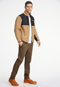 Chaqueta de forro polar con un cuerpo marrón claro y sección superior negra, que presenta un cuello alto y cremallera, combinada con pantalones marrones y botas de cordones marrones.