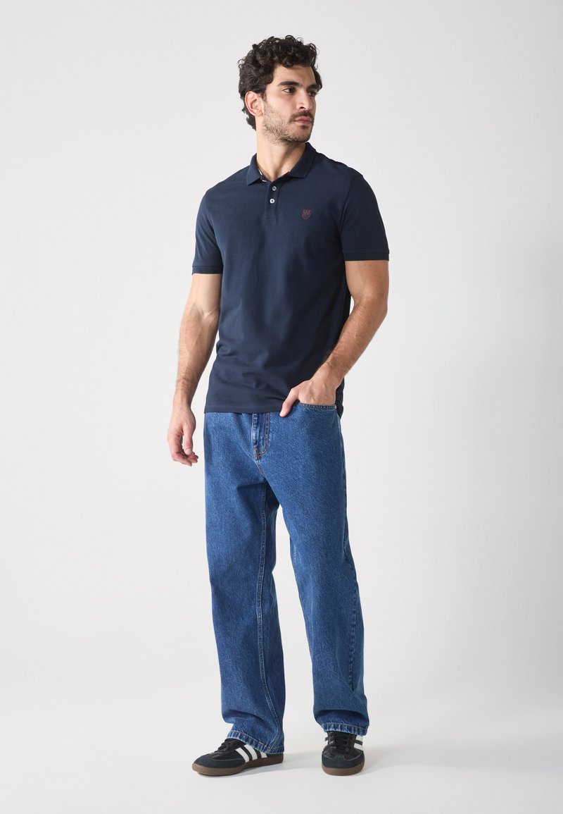 Polo de color marino, material de algodón, mangas cortas, con tapeta de botones en el frente y logo. Combinado con unos vaqueros de mezclilla azules holgados y zapatillas negras.