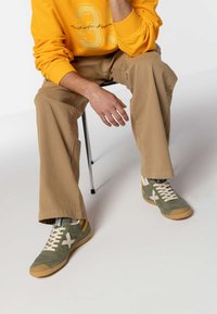 Sneakers verdi e grigi con una tomaia testurizzata, lacci beige e suola in gomma. Il modello indossa pantaloni khaki e una felpa gialla.