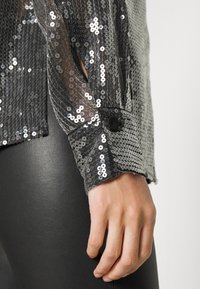 Blusa in paillettes argento con sovrapposizione trasparente, polsini con bottone nero e paillettes testurizzate, abbinata a leggings neri lisci.