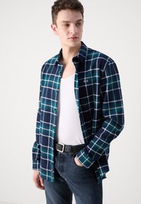 Camisa de franela a cuadros en azul marino, verde azulado y blanco, con un diseño de botones. Material de algodón, mangas largas y un pequeño logo en el pecho.