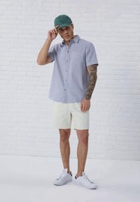 Striped short-sleeve shirt in blue and white, cream shorts, green cap, white sneakers; model has tattoos and stands against a white brick wall.