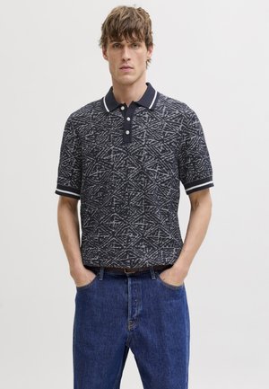 Mladi moški z razmršeno pričesko nosi črno-belo polo majico z geometrijskim vzorcem in modre jeans hlače, s rokami v žepih.