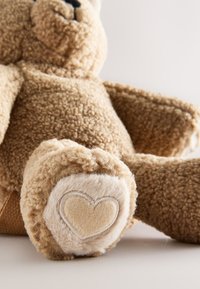 Blød brun teddybjørn med struktureret pels, sidder med en pote frem og viser et syet hjerte på fodpuden.