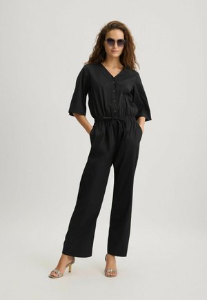 Zwarte jumpsuit met een v-hals, korte mouwen en een koordtaille. Gekenmerkt door knopen en wijde broekspijpen. Draagt met metallic haksandalen.